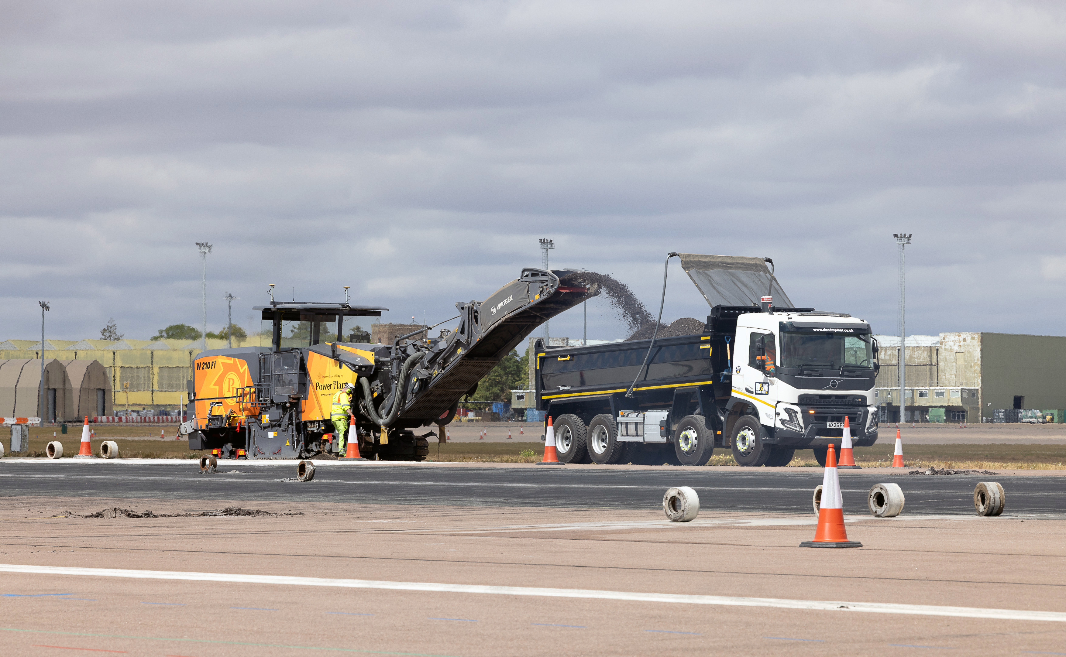 A yellow and black Wirtgen road milling machine discharges excavated asphalt via a conveyor belt into a large white Volvo tipper truck. The construction vehicles are actively operating on an airport runway under an overcast sky, surrounded by traffic cones.